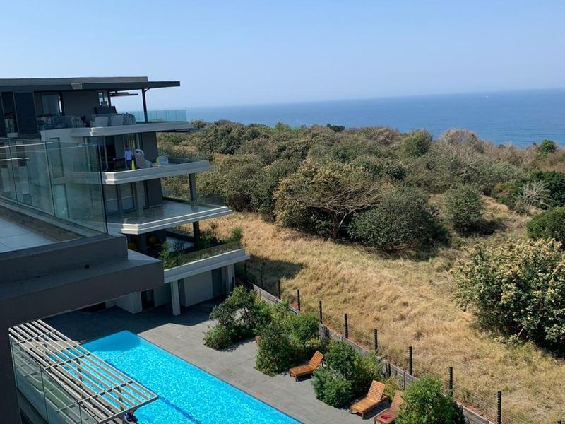 Elevated view of a modern apartment complex featuring a long lap pool, glass-railed balconies, and scenic coastal views.