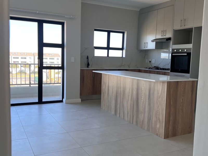 Modern open-plan kitchen featuring a central island, built-in oven, extractor fan, stone countertops, and direct access to a balcony through sliding glass doors.