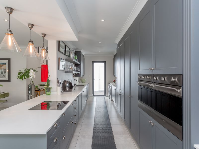 Modern galley-style kitchen featuring dark grey cabinetry, white quartz countertops, integrated appliances, and sleek pendant lighting, leading to a rear exterior door.