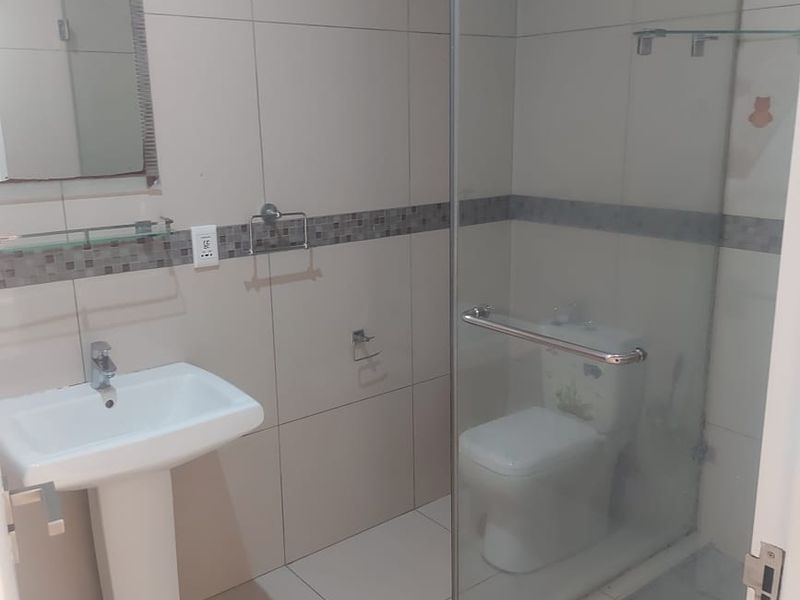 A clean, modern bathroom featuring a pedestal washbasin, a glass-enclosed shower, and a toilet with neutral wall and floor tiling.