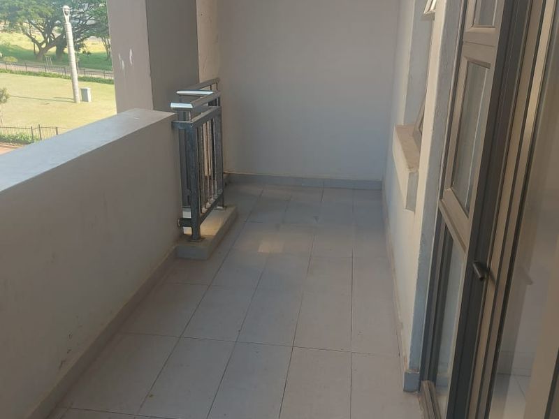 A clean, empty balcony featuring tiled flooring, a metal balustrade, and sliding glass door access, offering a view of the surrounding estate grounds.