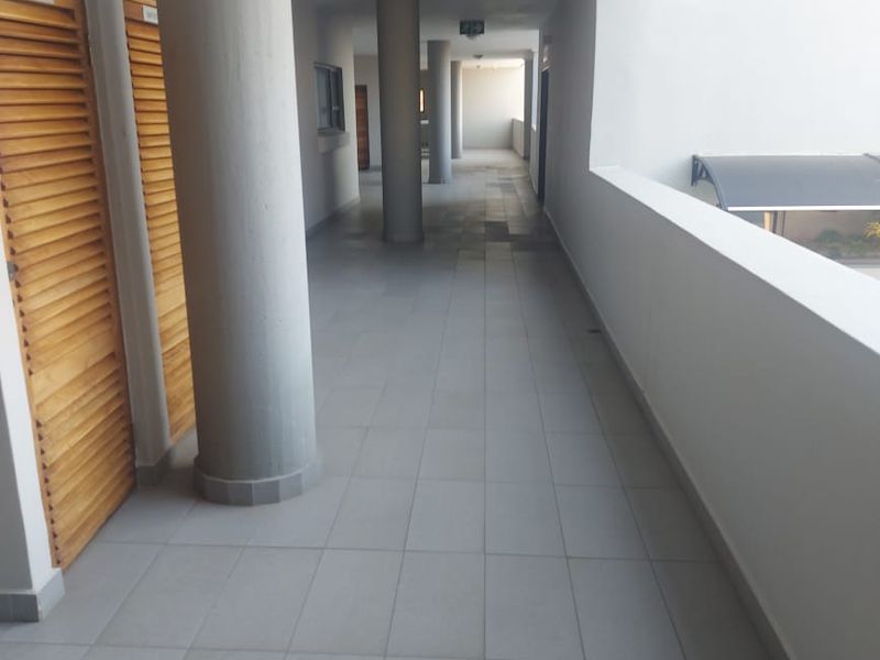 A clean, open-air apartment hallway featuring tiled flooring, structural columns, and louvered utility access doors.