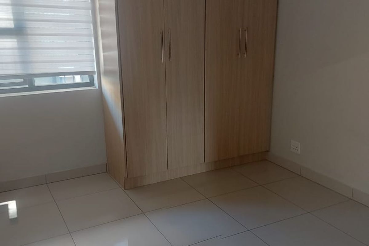 A clean, empty bedroom featuring light-toned built-in wardrobes, neutral tiled flooring, and a modern roller blind on the window.