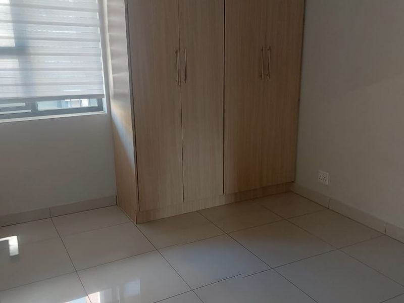 A clean, empty bedroom featuring light-toned built-in wardrobes, neutral tiled flooring, and a modern roller blind on the window.