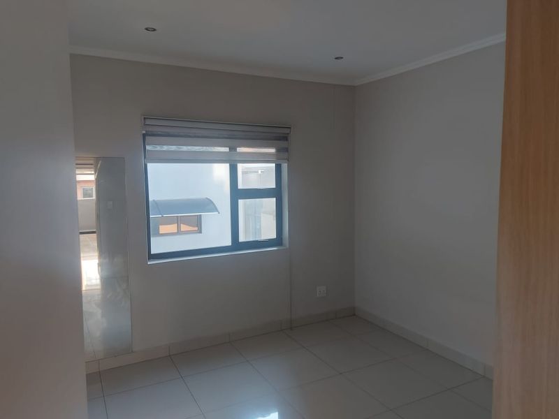 A clean, empty bedroom featuring neutral tiled flooring, recessed downlights, and an aluminium-framed window with a fitted roller blind.