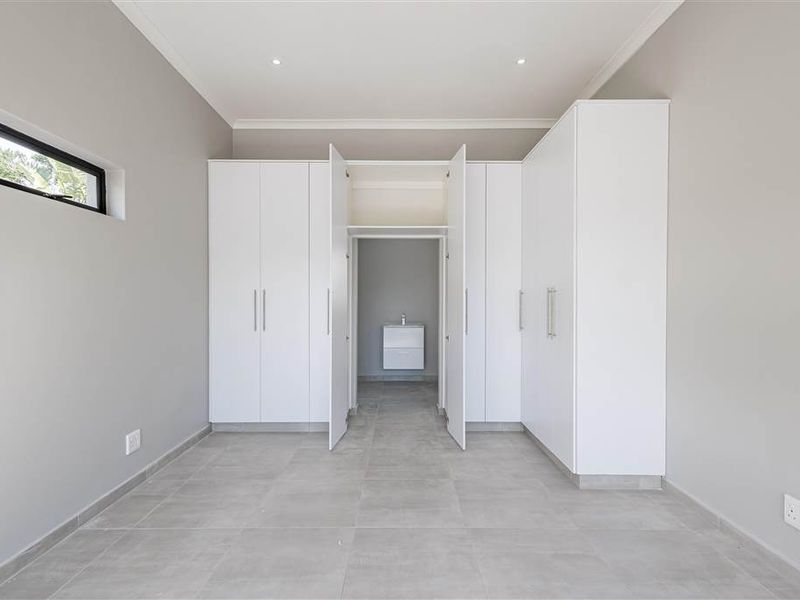 A modern, minimalist bedroom featuring extensive built-in cabinetry, neutral tiled flooring, and a private en-suite vanity area.