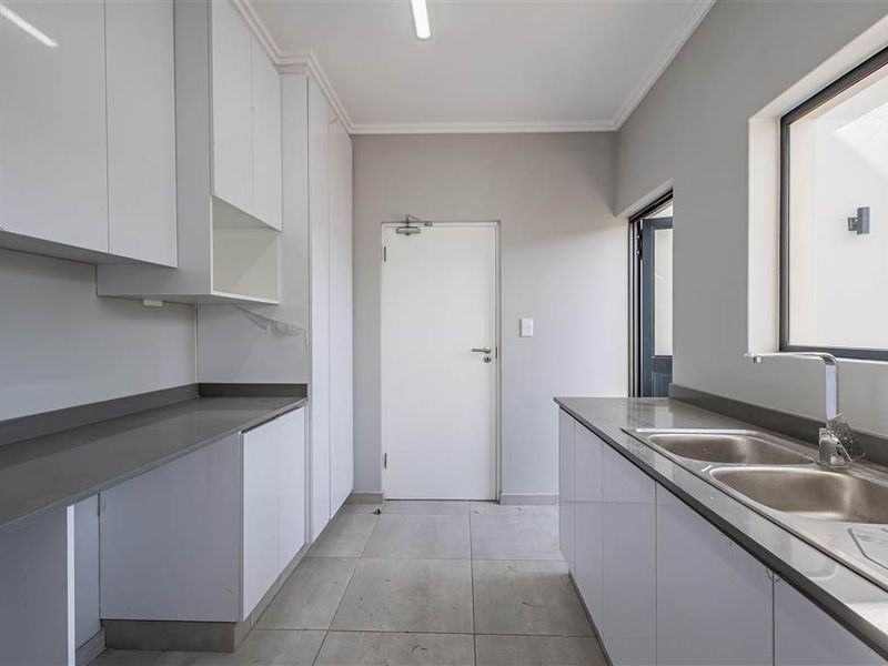 A clean and functional scullery featuring modern white cabinetry, stone countertops, a double bowl sink, and tiled flooring.