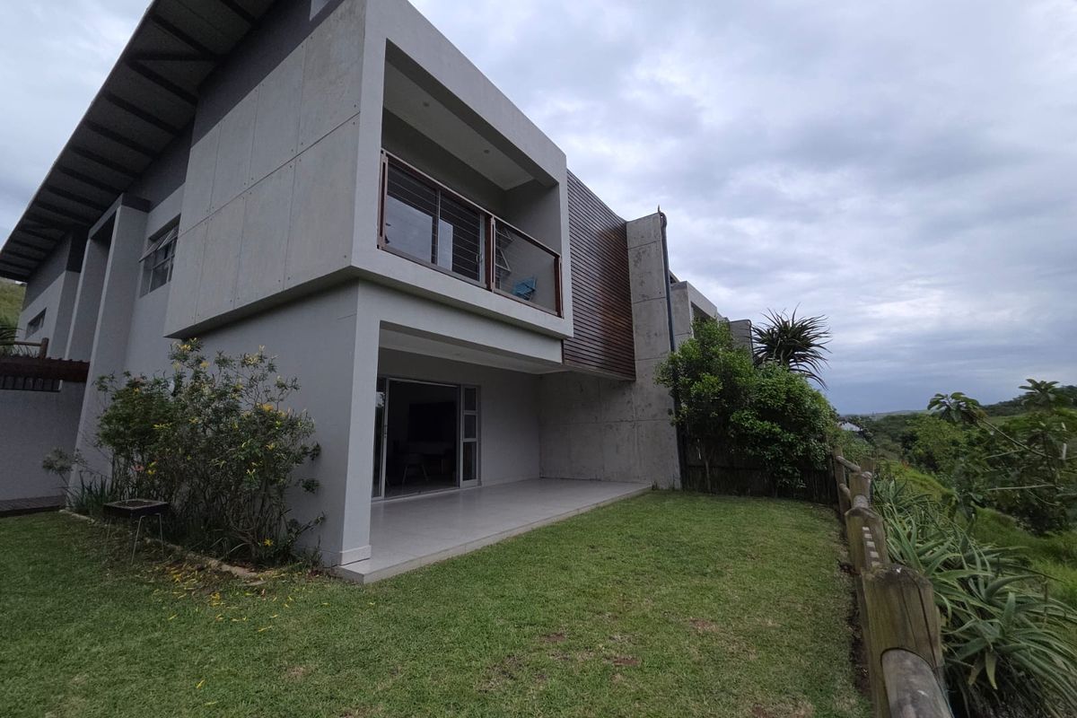 Rear view of a modern two-story house featuring concrete and timber cladding, a covered patio, and a balcony with glass balustrades overlooking the garden.