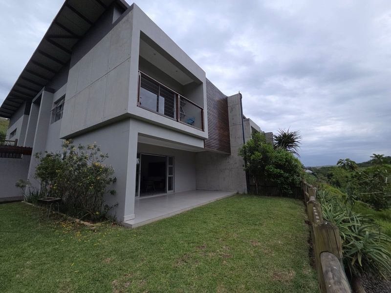 Rear view of a modern two-story house featuring concrete and timber cladding, a covered patio, and a balcony with glass balustrades overlooking the garden.