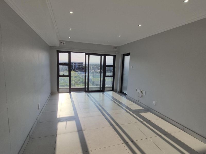 A bright, empty living area featuring neutral tiled flooring, recessed downlighting, and large sliding glass doors leading to an outdoor balcony.
