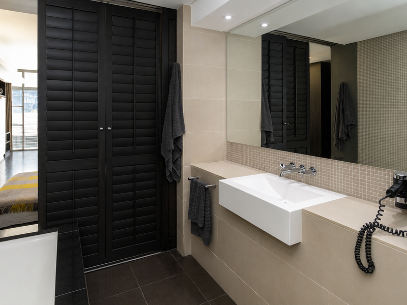 Modern bathroom featuring a wall-mounted basin, large vanity mirror, mosaic tile splashback, and dark shutter-style cabinetry, with a view into the adjacent living area.