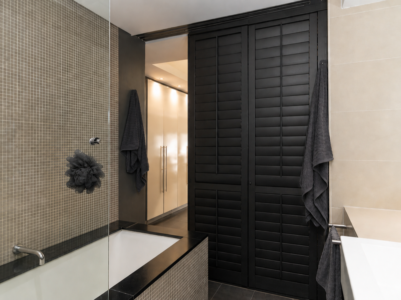 Modern bathroom featuring a built-in bathtub with mosaic tiling, a glass screen, and elegant black plantation shutters providing privacy and a contemporary aesthetic.