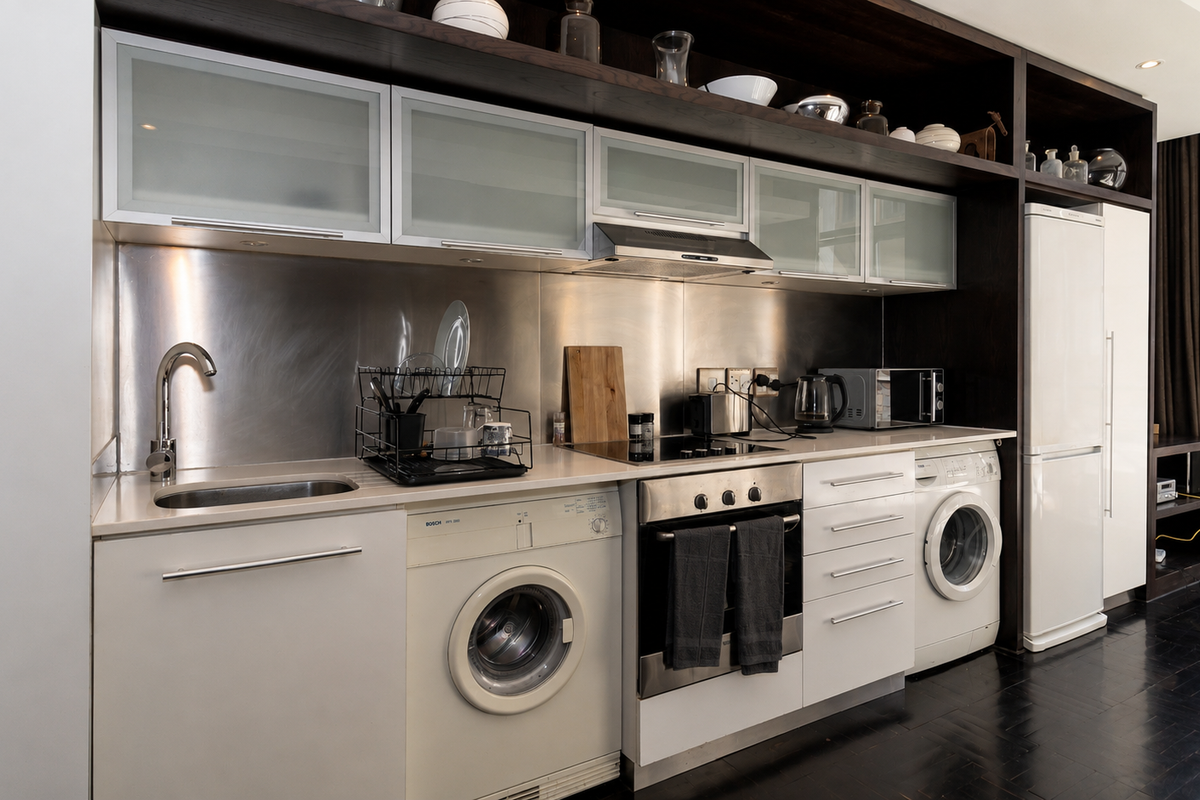 Modern compact kitchen featuring stainless steel splashbacks, integrated appliances including oven, hob, and laundry units, with dark wood cabinetry and open shelving.