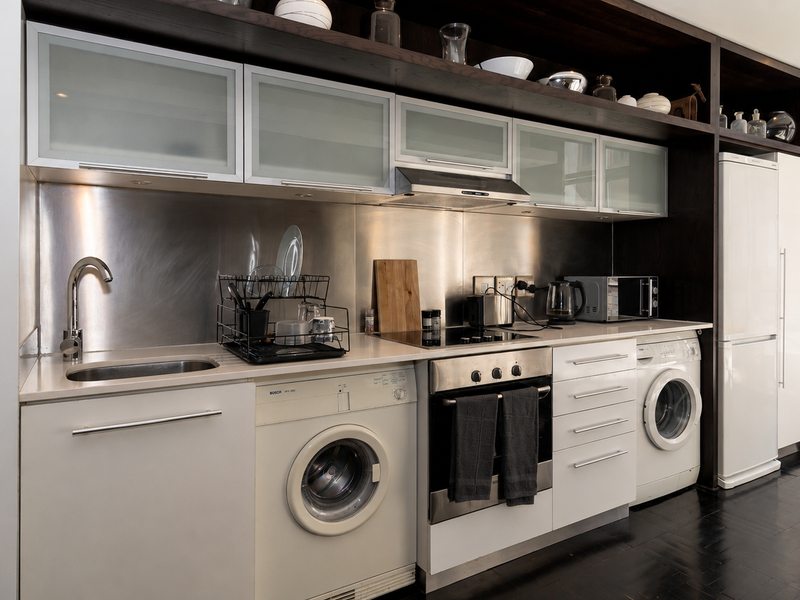 Modern compact kitchen featuring stainless steel splashbacks, integrated appliances including oven, hob, and laundry units, with dark wood cabinetry and open shelving.