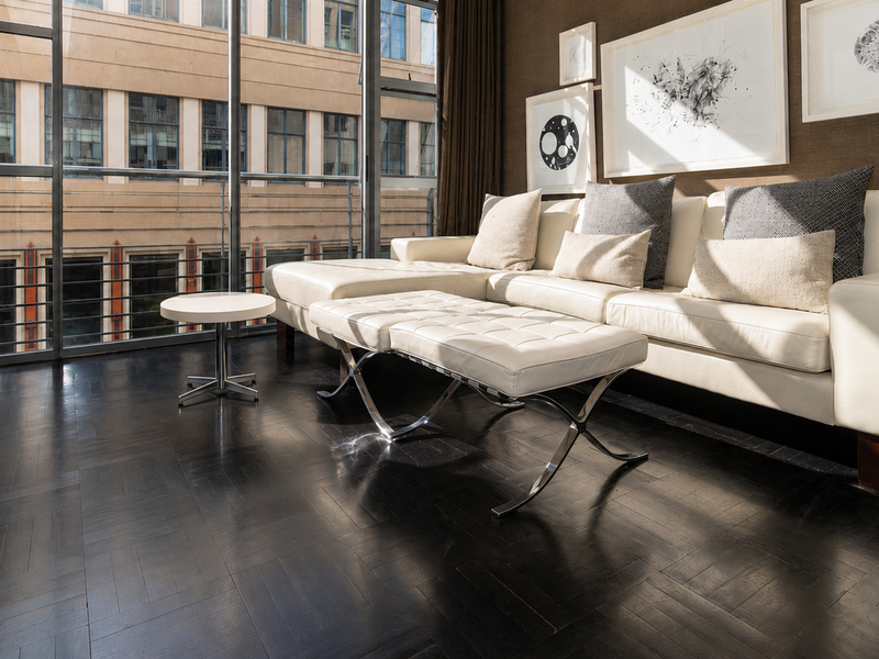 A modern living room featuring dark parquet flooring and large floor-to-ceiling windows that provide ample natural light and an urban view.