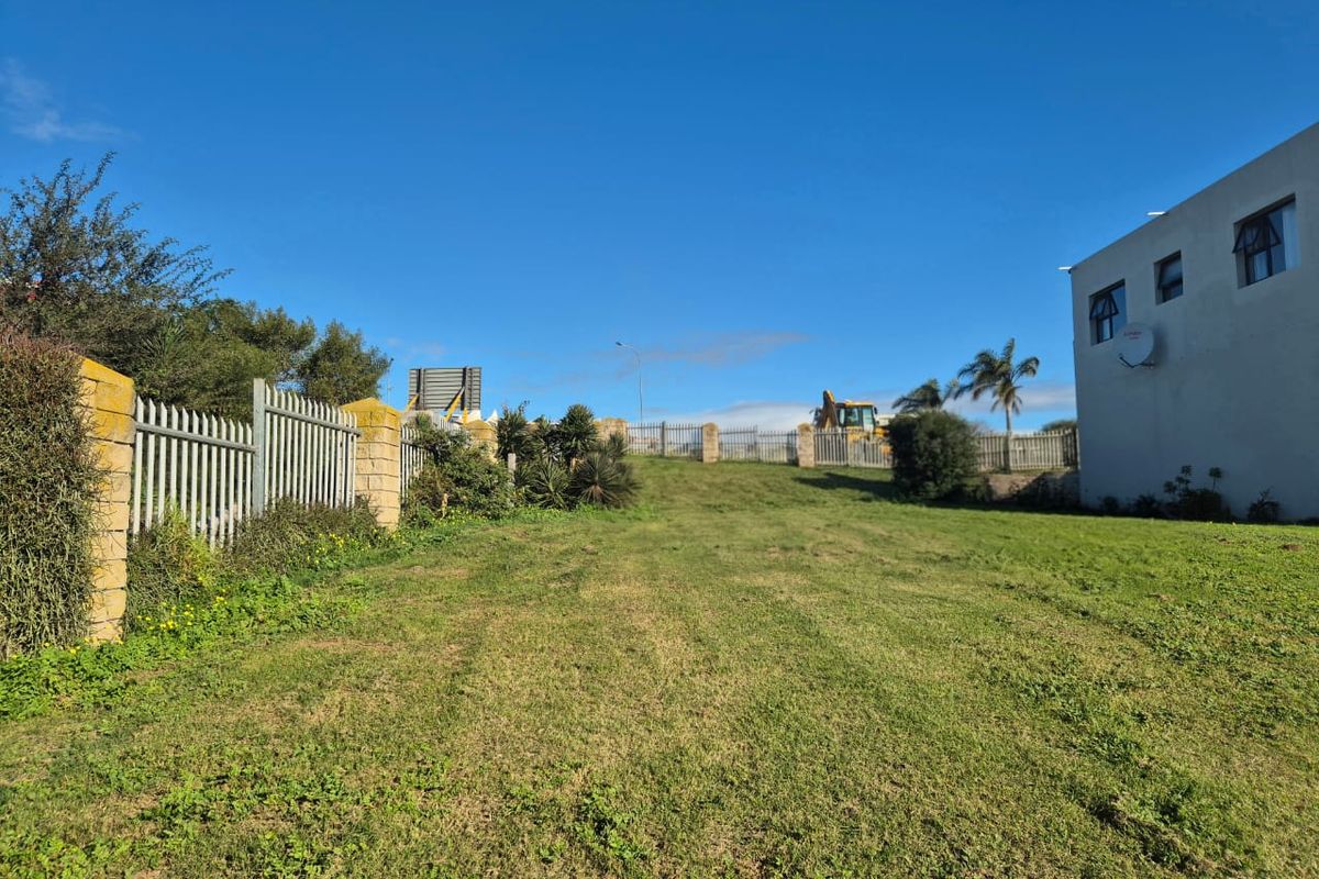 A view of a residential vacant land plot featuring a grassy lawn, metal palisade fencing, and stone masonry pillars.
