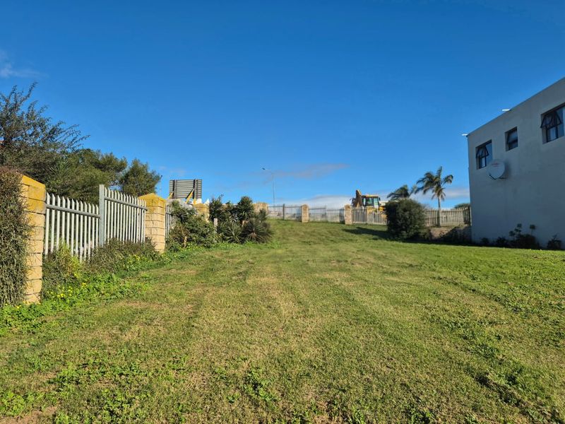 A view of a residential vacant land plot featuring a grassy lawn, metal palisade fencing, and stone masonry pillars.