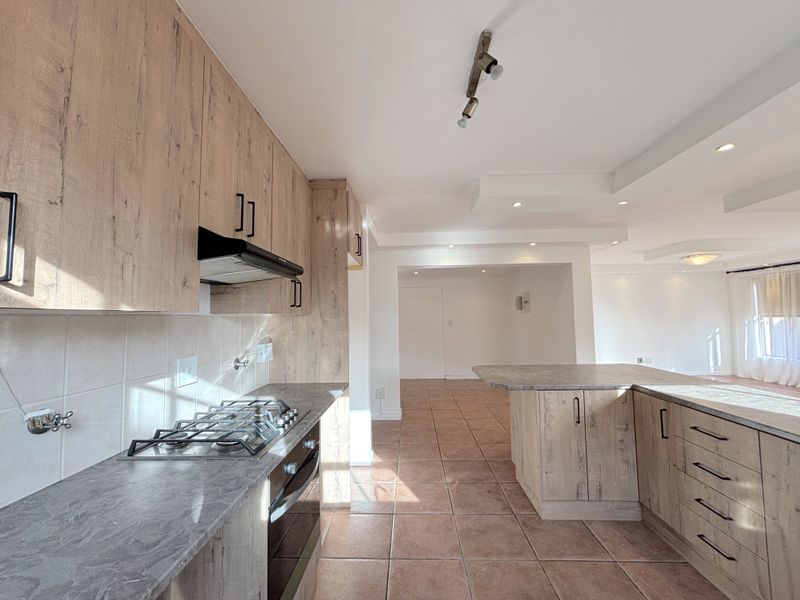 Open-plan kitchen featuring wood-look cabinetry, stone-effect countertops, gas hob, and integrated oven, flowing into a spacious living area with tiled flooring.