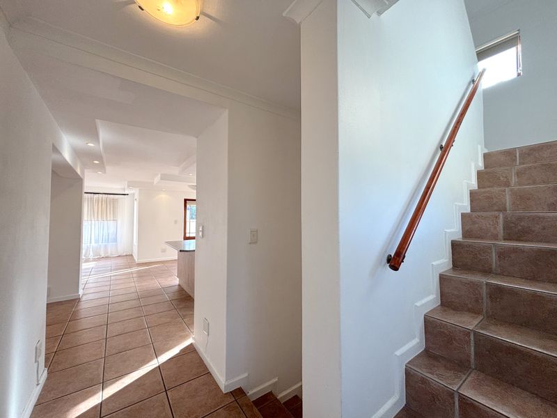 A bright, tiled hallway featuring a staircase with a wooden handrail, leading towards an open-plan living area with recessed ceiling lighting.
