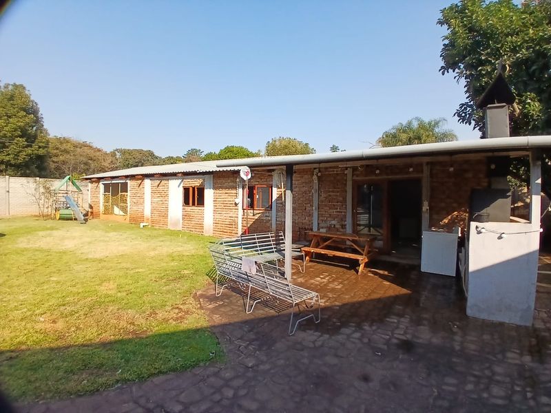 Rear view of the property featuring an exposed brick facade, a covered patio area with a built-in braai, and a spacious garden.
