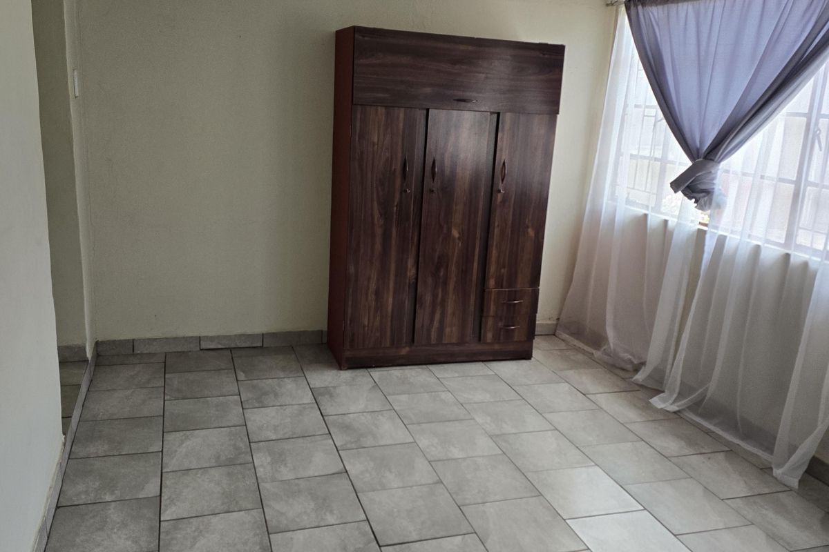A simple, tiled bedroom featuring neutral walls, a ceiling light fixture, and a window with curtains.