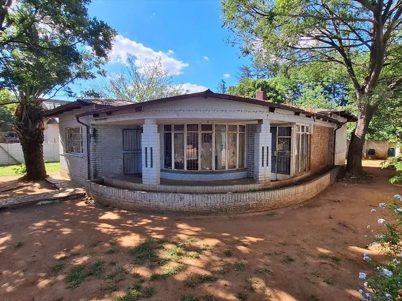 Rear view of a single-story brick house featuring a curved veranda, metal-framed windows, and a large, unlandscaped garden area.