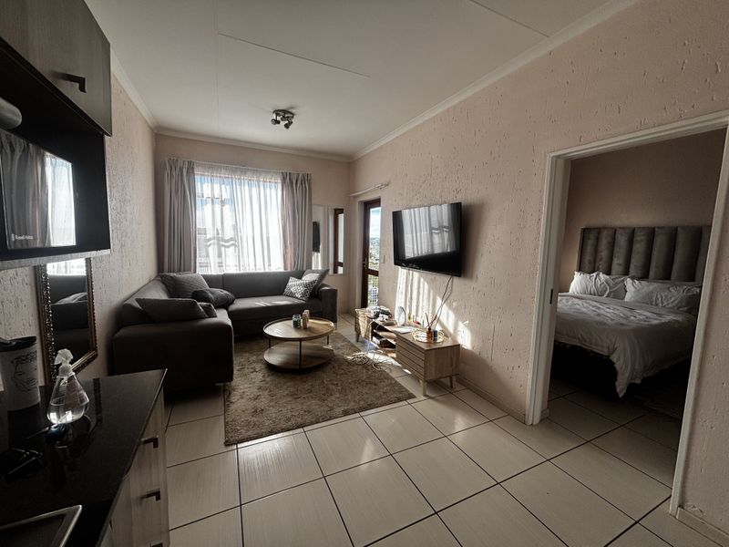 A living room featuring tiled flooring and textured walls, with direct access to an adjacent bedroom and natural light from the balcony door.