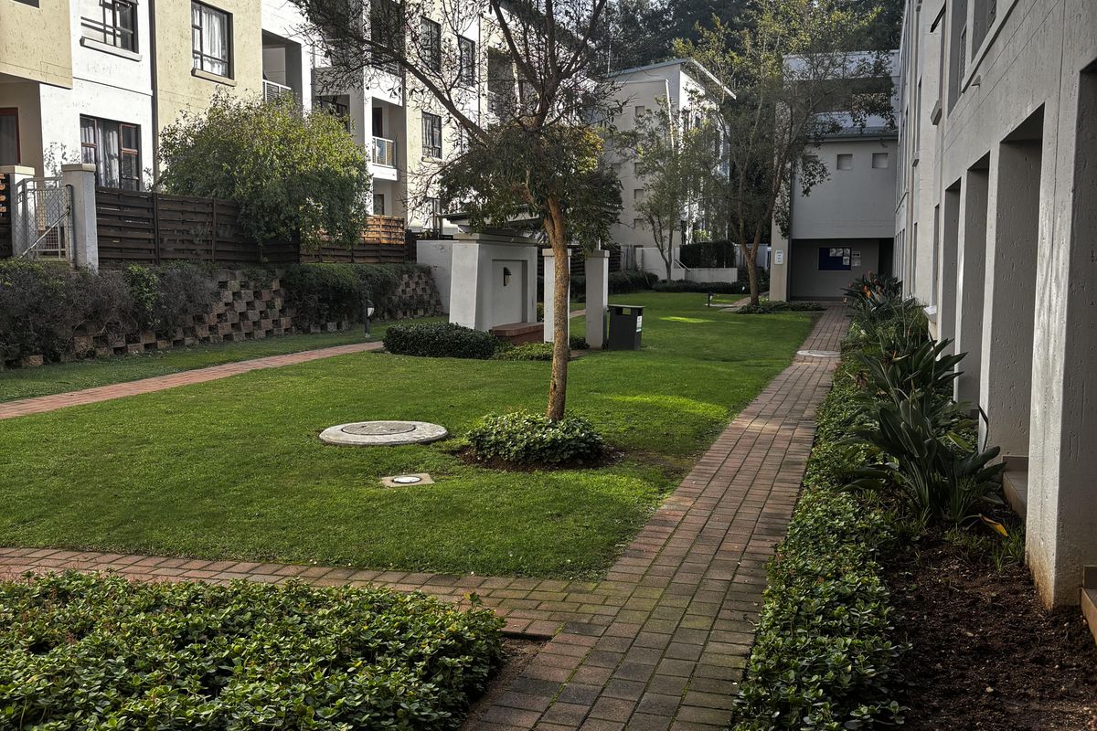 A well-maintained communal garden area between apartment blocks, featuring paved walkways, manicured lawns, and established landscaping.