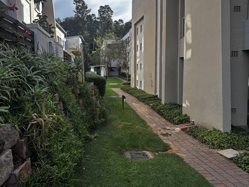 A view of the exterior rear grounds featuring a brick paved pathway, landscaped garden beds, and a retaining wall between apartment blocks.
