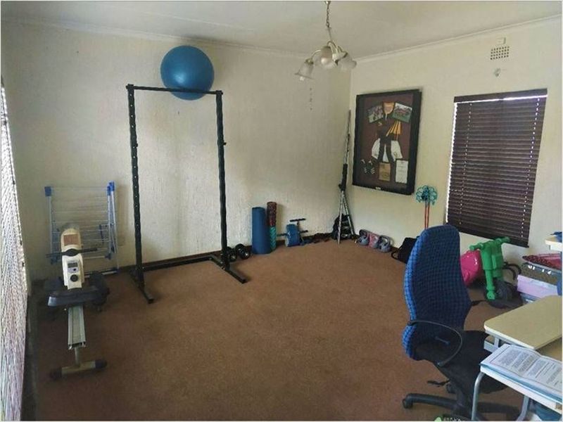 A multi-purpose room featuring brown carpeting, venetian blinds, and a central ceiling light fixture, currently utilized as a home gym and office space.