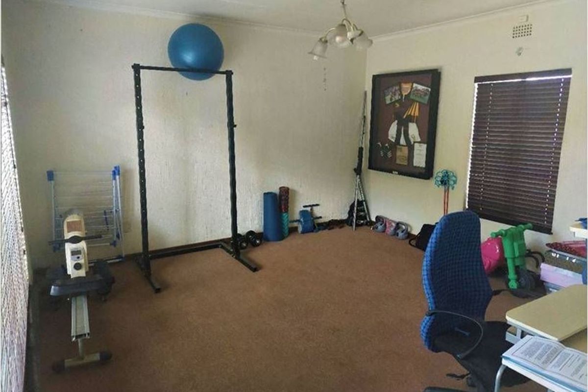 A multi-purpose room featuring brown carpeting, venetian blinds, and a central ceiling light fixture, currently utilized as a home gym and office space.