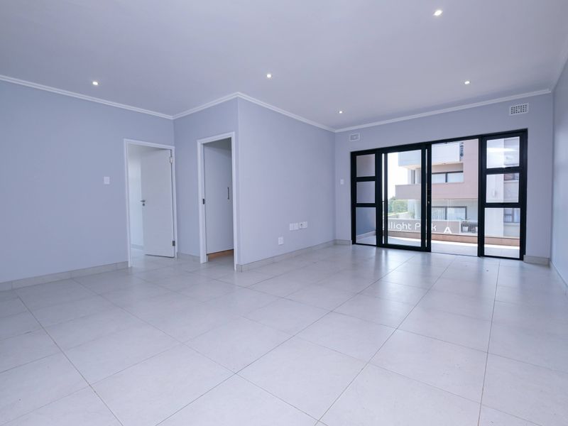 A bright, spacious, and empty living area featuring neutral tiled flooring, recessed lighting, and large sliding glass doors leading to an outdoor balcony.