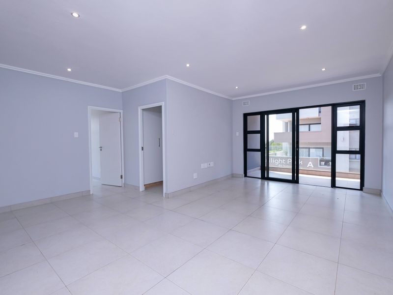 A bright, empty living room featuring neutral tiled flooring, recessed downlights, and large sliding glass doors leading to an outdoor balcony area.