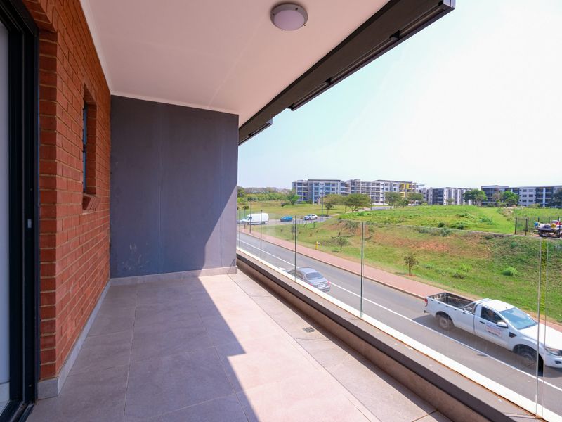 A spacious covered balcony featuring modern glass balustrades, tiled flooring, and an exposed brick feature wall, offering an open outlook over the surrounding area.