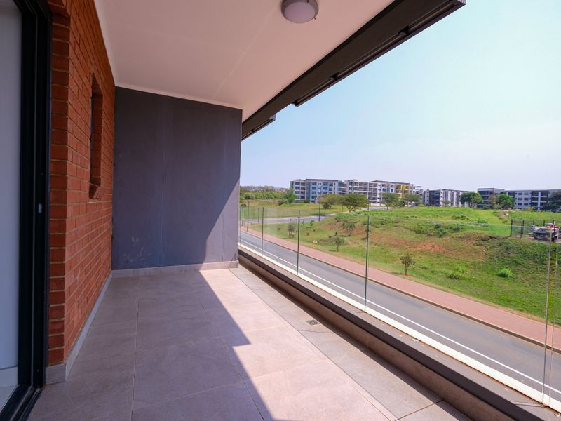A spacious covered balcony featuring modern tiled flooring, an exposed brick feature wall, and a clear glass balustrade overlooking the surrounding neighbourhood.