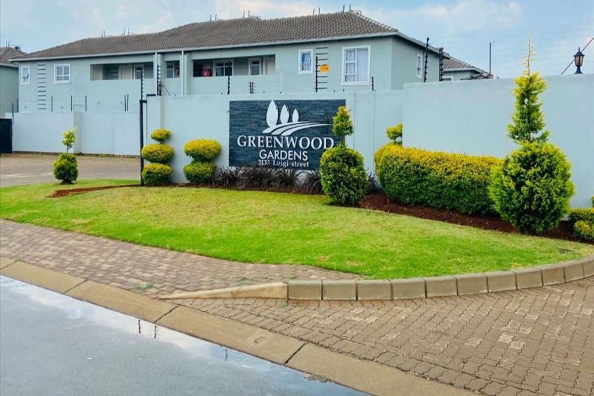 Front entrance view of Greenwood Gardens apartment complex featuring a paved driveway, landscaped garden, and secure perimeter wall with electric fencing.