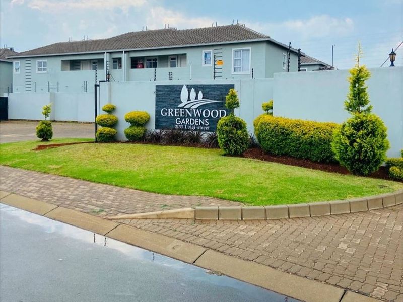 Front entrance view of Greenwood Gardens apartment complex featuring a paved driveway, landscaped garden, and secure perimeter wall with electric fencing.