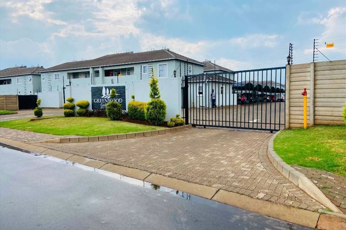 Front entrance view of Greenwood Gardens apartment complex featuring a paved driveway, secure gated access, and perimeter wall with electric fencing.