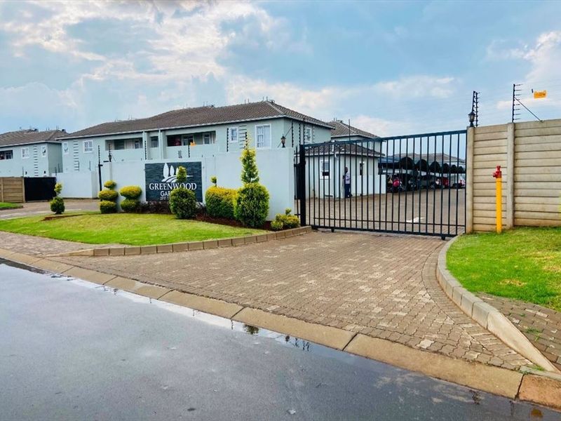 Front entrance view of Greenwood Gardens apartment complex featuring a paved driveway, secure gated access, and perimeter wall with electric fencing.