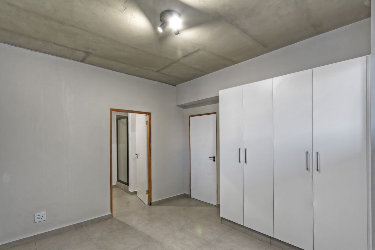 A clean, modern bedroom featuring built-in white cupboards, tiled flooring, and an exposed concrete ceiling, offering a neutral canvas for personalization.