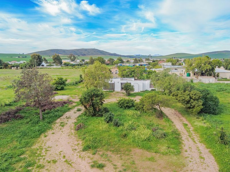 Aerial view of a residential property featuring a large plot of land with mature trees and an unpaved driveway approach.