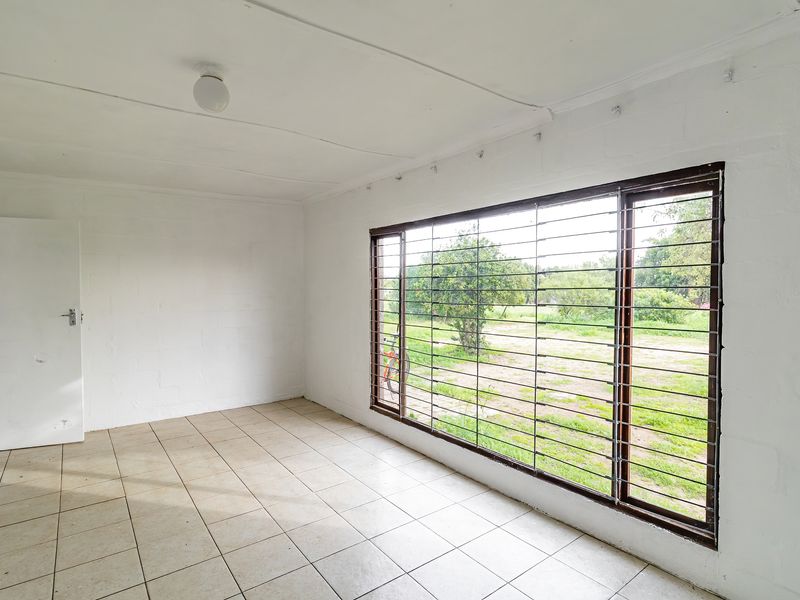 An empty living room featuring tiled flooring, painted blockwork walls, and a large window fitted with security bars overlooking the garden.