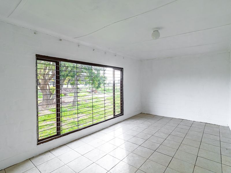 An empty, sunlit living area featuring tiled flooring, a large window with security bars, and a neutral white finish throughout.