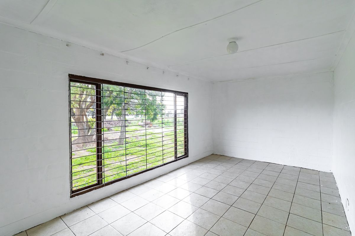 An empty, sunlit living area featuring tiled flooring, a large window with security bars, and a neutral white finish throughout.