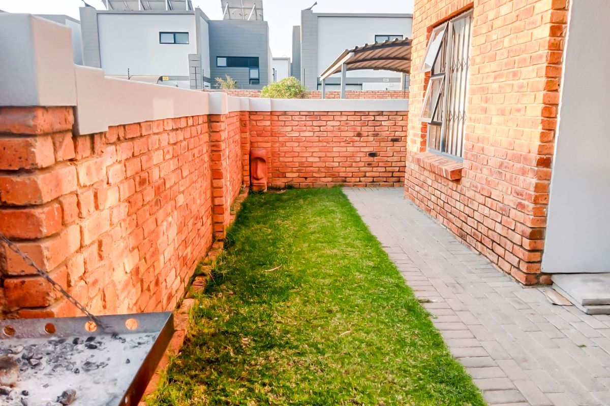 A private rear garden area featuring a paved walkway, a small lawn, and a built-in braai against a face brick boundary wall.