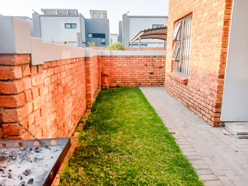 A private rear garden area featuring a paved walkway, a small lawn, and a built-in braai against a face brick boundary wall.