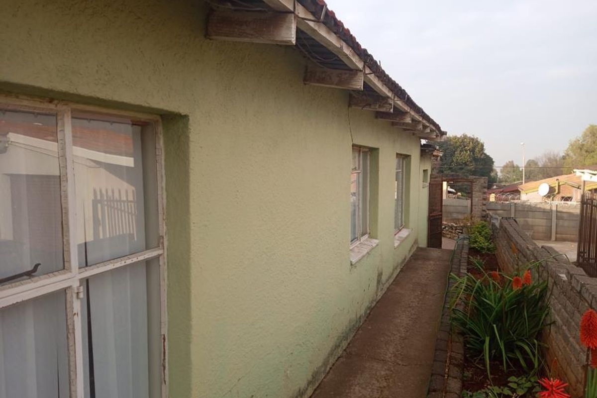 A side view of the property showing a concrete pathway, plastered exterior walls, and exposed timber roof rafters along the building perimeter.