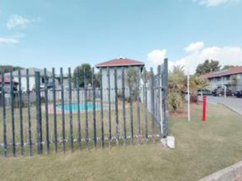Communal swimming pool area featuring a paved surround and metal security fencing within a landscaped residential complex setting.