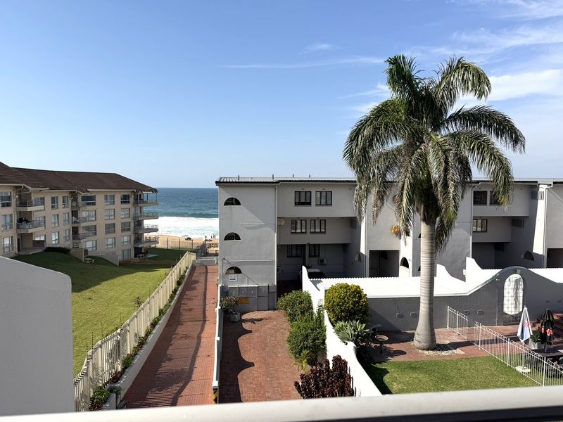 Elevated view overlooking the apartment complex grounds, paved driveway, landscaped garden, and the ocean coastline in the distance.