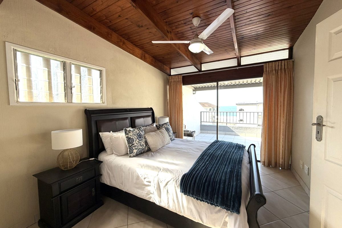 Spacious bedroom featuring a vaulted timber ceiling, ceiling fan, tiled floors, and direct access to a private balcony with ocean views.
